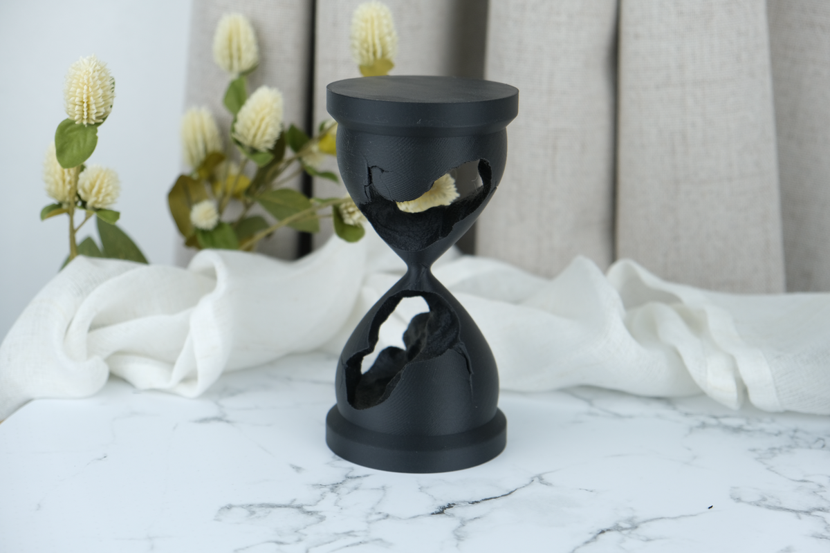 Skulptur av ett trasigt timglas på en mörk hylla med böcker och abstrakta föremål