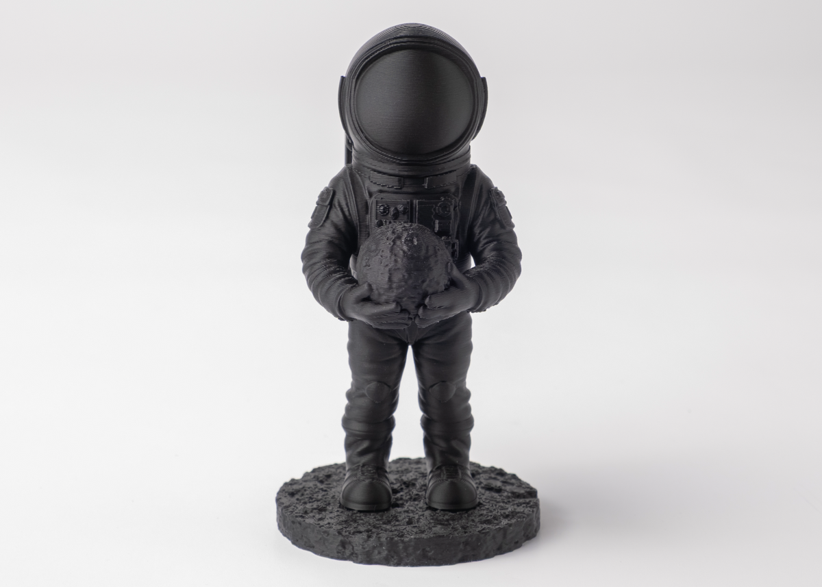 Mörk astronautskulptur på en trähylla bredvid böcker och en liten växt