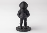 Mörk astronautskulptur på en trähylla bredvid böcker och en liten växt