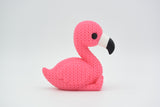 Stickad flamingofigur på en trähylla bredvid en liten växt och neutral inredning